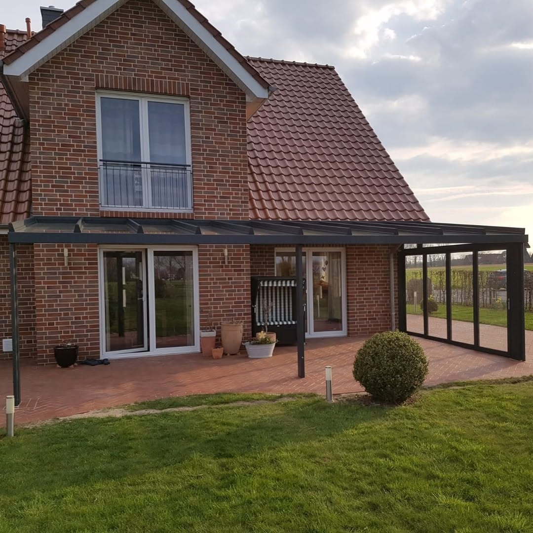 Einfamilienhaus mit roter Klinkerfassade und Satteldach, davor eine schwarze Terrassenüberdachung mit seitlicher Verglasung und angrenzendem Garten.