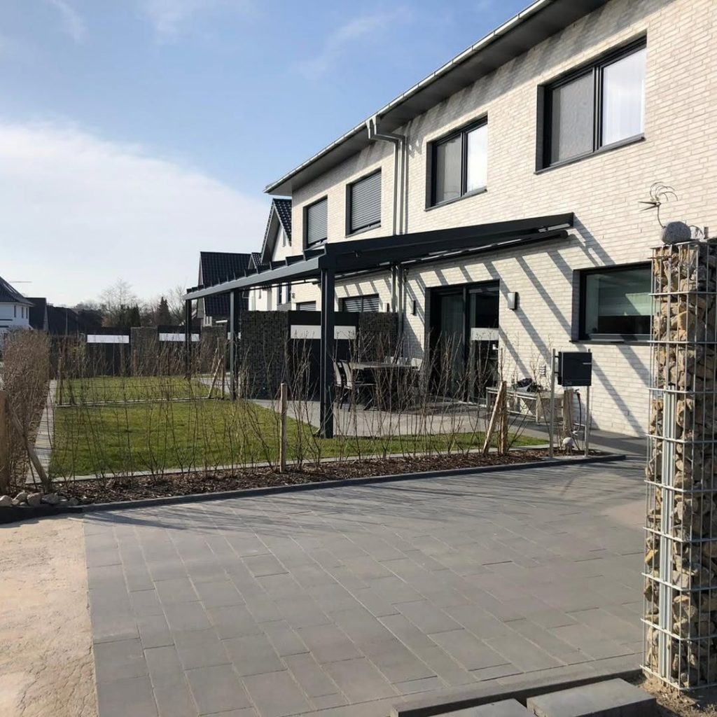Reihenhäuser mit heller Klinkerfassade und moderner Terrassenüberdachung aus Metall im Gartenbereich, aufgenommen bei sonnigem Wetter.
