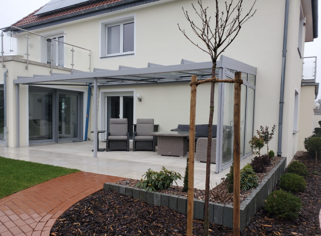Moderne Terrassenüberdachung aus Aluminium an einem weißen Einfamilienhaus, mit Sitzgruppe auf der Terrasse und neu angelegtem Gartenbereich.