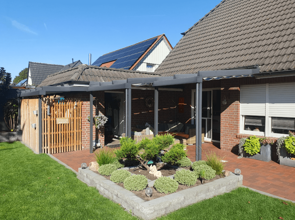 Aluminium-Terrassenüberdachung im Garten neben einem Wohnhaus, umgeben von Grünflächen, Pflanzen und dekorativer Gartengestaltung
