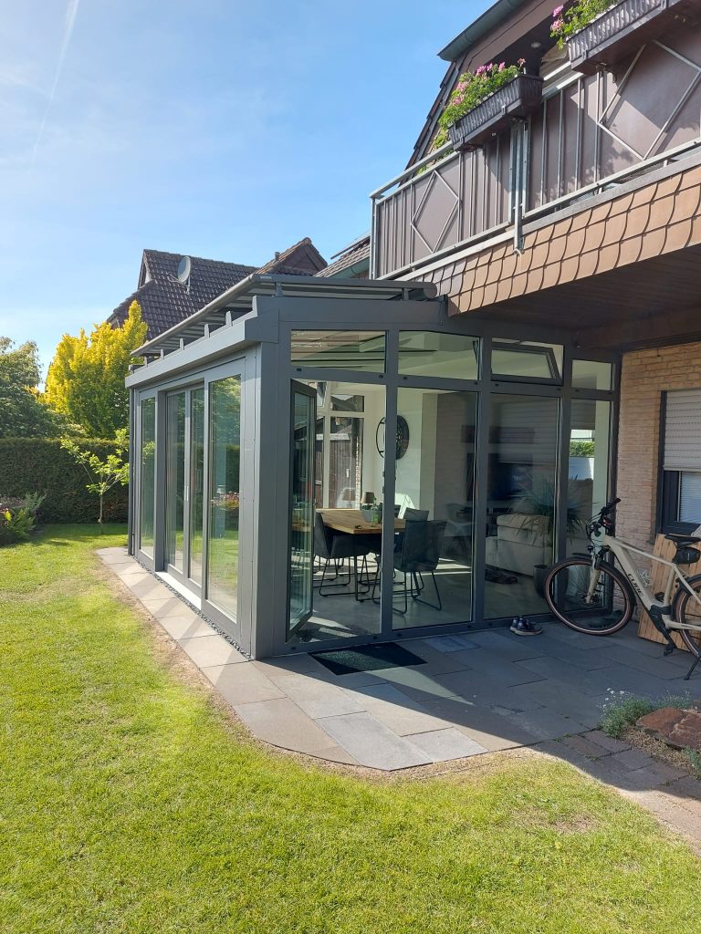 Warmer Wintergarten mit grauem Aluminiumrahmen und großen Glasfronten an einem Einfamilienhaus, genutzt als heller Wohn- und Essbereich.