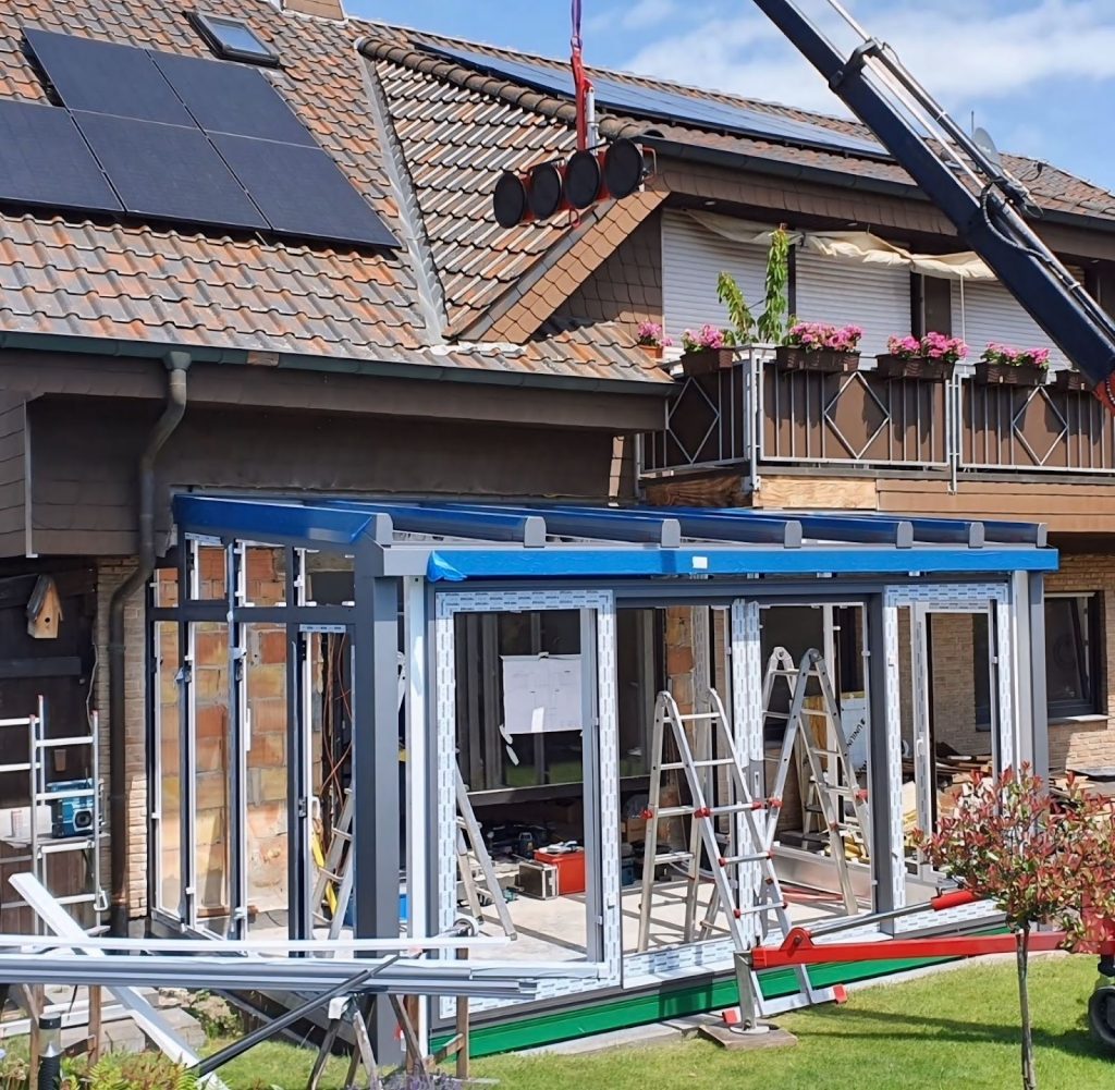 Montage eines Wintergartens mit Aluminiumrahmen und Glasdach, bei dem Bauelemente per Kran auf das Wintergarten Dach gehoben werden.